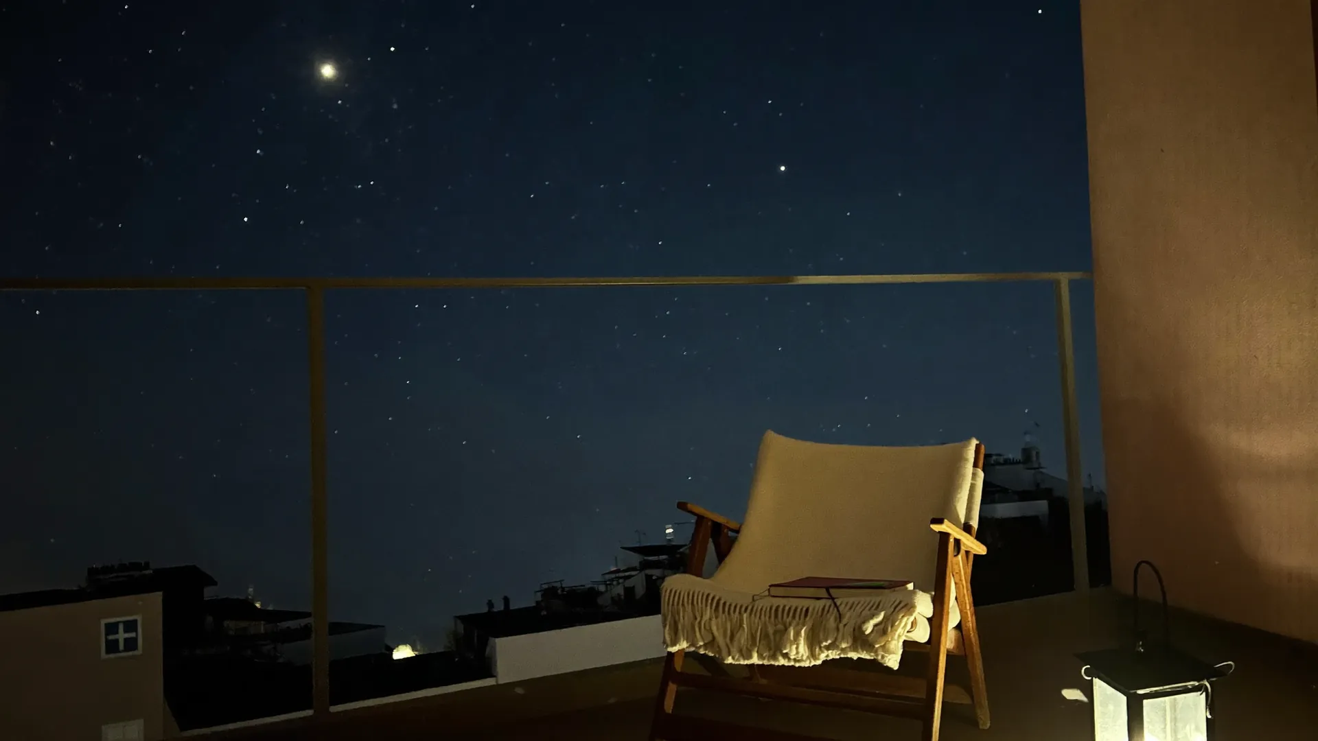 Ein leerer Balkon bei Nacht, auf dem Stuhl liegen eine Decke und ein zugeklapptes Notizbuch, daneben leuchtet eine Laterne, darüber ein heller Stern