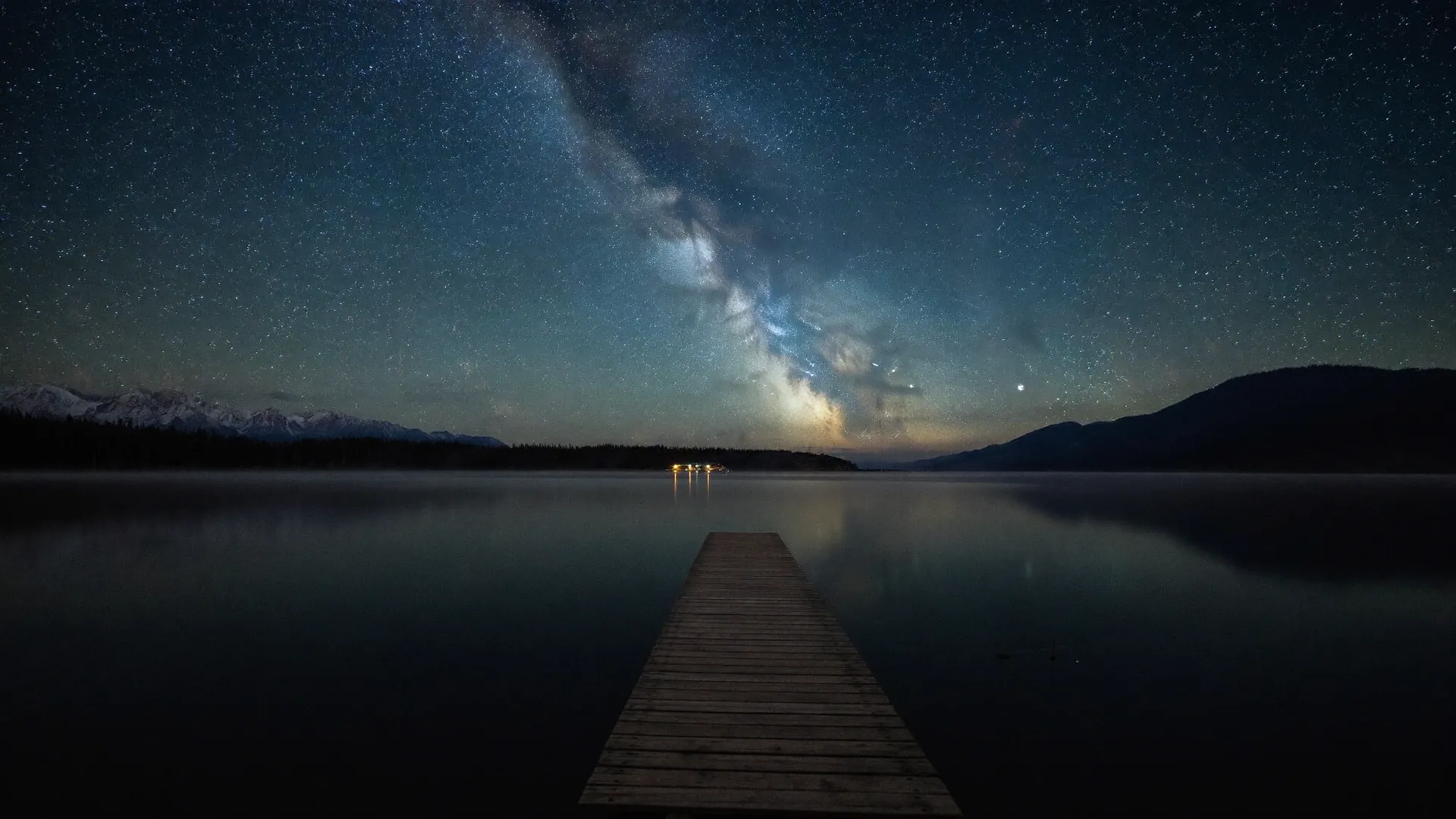 Ein stiller Bergsee bei Nacht mit einem Holzsteg, darüber ein klarer Sternenhimmel mit der Milchstraße, der sich im Wasser spiegelt
